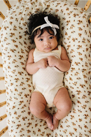 A baby girl in a light yellow pointelle tank bodysuit rests on a floral-patterned cushion, wearing a white bow headband.