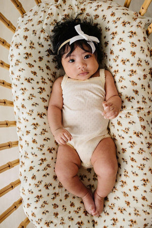 A baby girl lies on a floral-patterned blanket, wearing a light yellow pointelle tank bodysuit and a white hair bow.