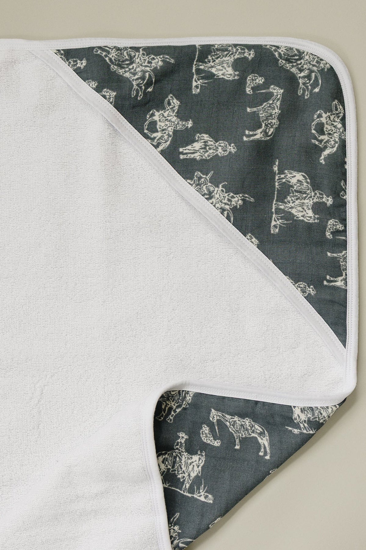 White muslin hooded towel with a dark gray western pattern, featuring cowboys and horses, on a beige background.