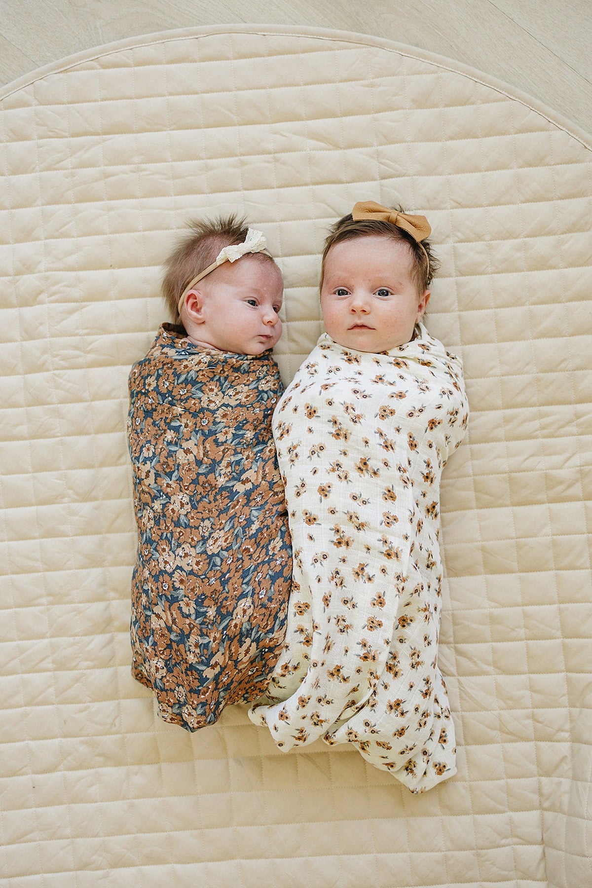 Two babies wrapped in floral muslin swaddle blankets on a quilted beige mat, wearing matching headbands.