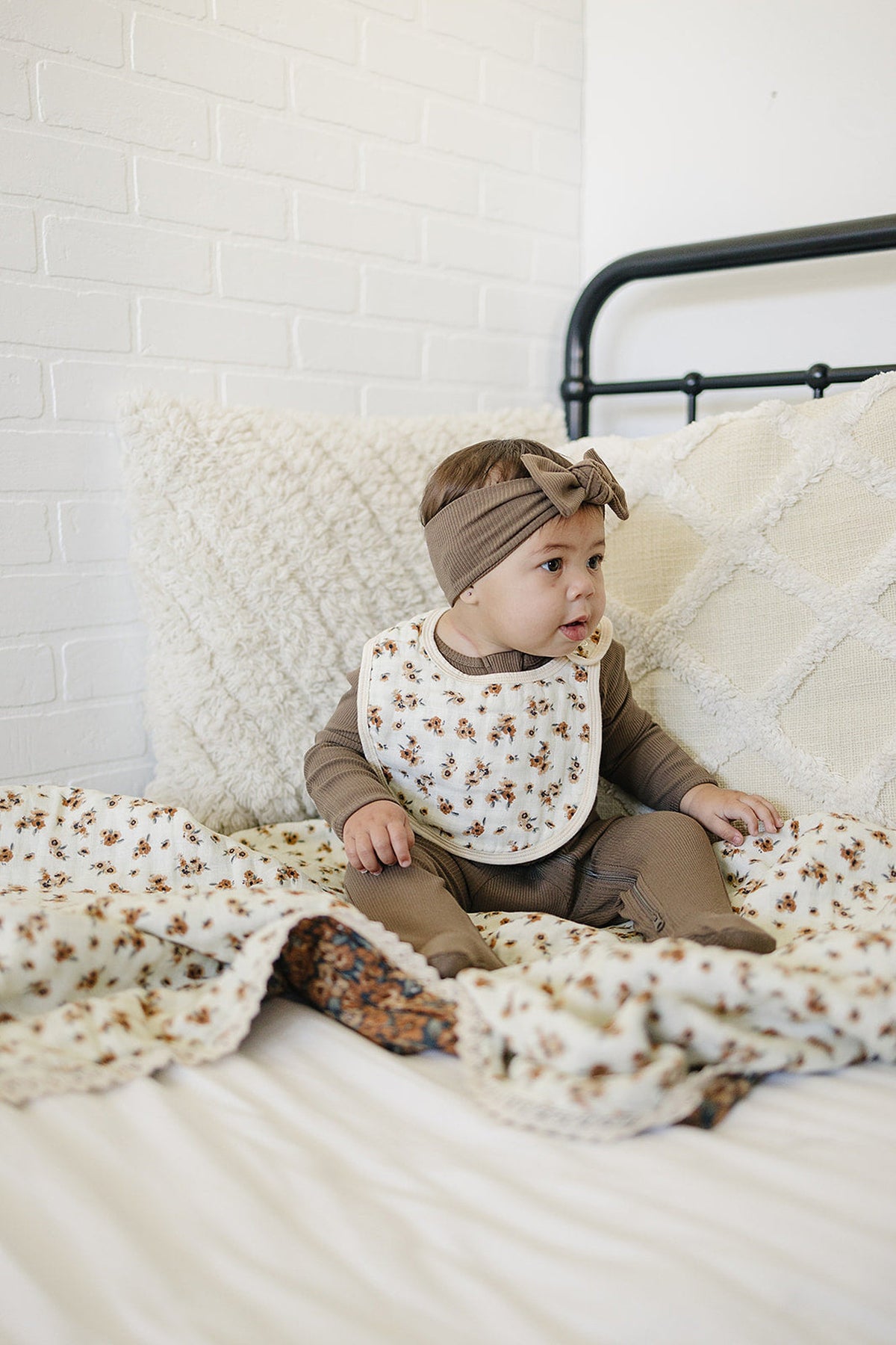A baby wearing a floral bib and brown outfit sits on a bed with a floral quilt.