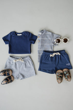 A navy ribbed t-shirt and a grey striped button-up are paired with matching shorts and sandals, arranged on a light.