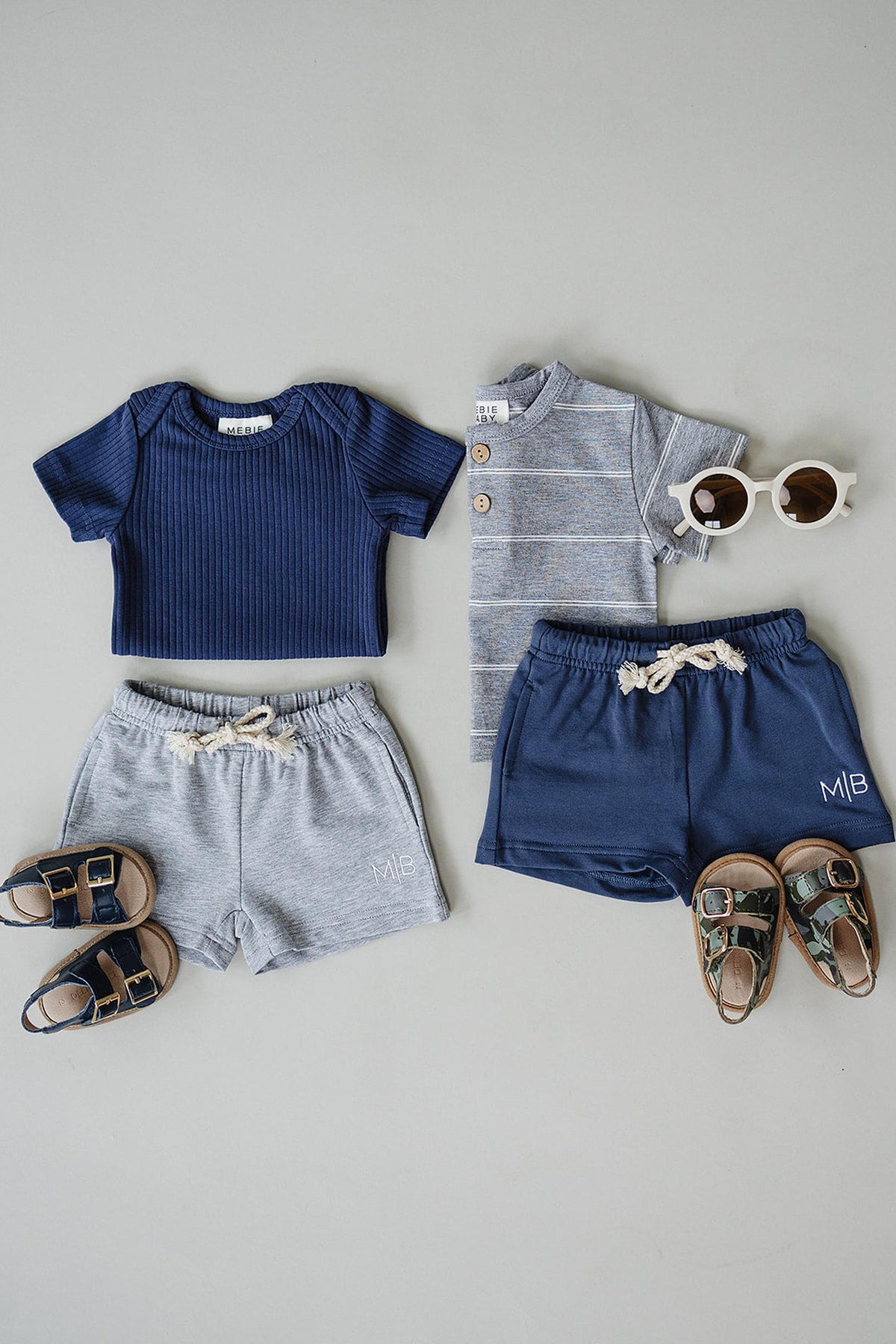 A flat lay of childrens clothing features a navy ribbed shirt, a striped grey button-up, grey cotton shorts, and navy shorts, paired with stylish sunglasses and sandals.