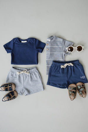 A flat lay of childrens clothing features a navy ribbed shirt, a striped grey button-up, grey cotton shorts, and navy shorts, paired with stylish sunglasses and sandals.