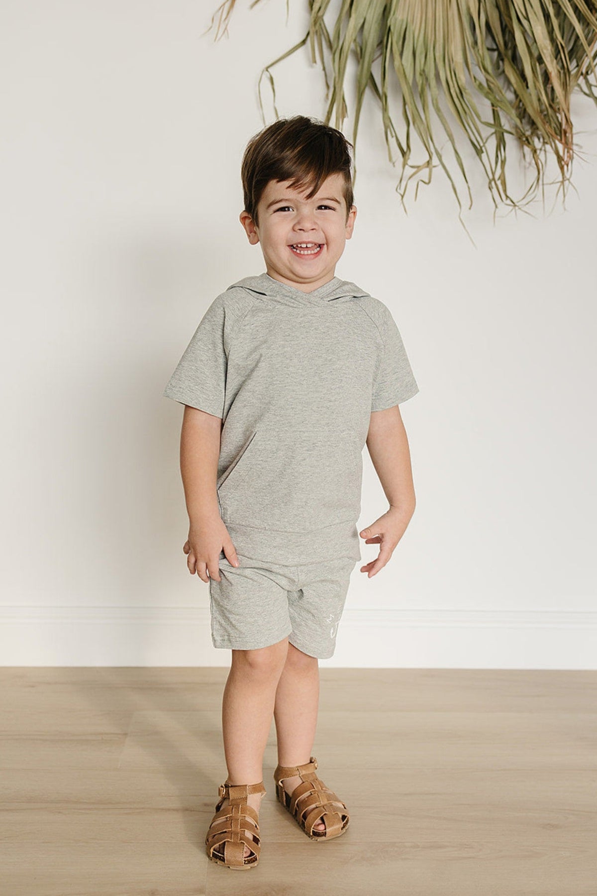 A smiling toddler wears a heather grey hooded t-shirt and matching shorts, paired with brown sandals, standing on a light.