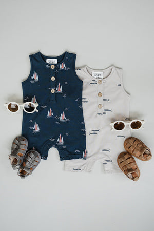 Two cotton short rompers for baby boys are displayed, one navy with sailboat print and one light beige with fish design.