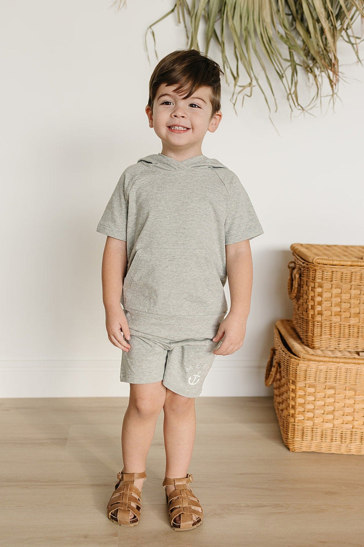 A smiling child wears a heather grey matching set with shorts, standing near wicker baskets on a wooden floor.