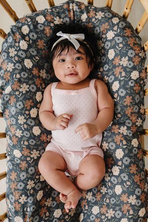 A baby girl in a pink pointelle tank bodysuit sits on a floral-patterned cushion, smiling with a white bow in.