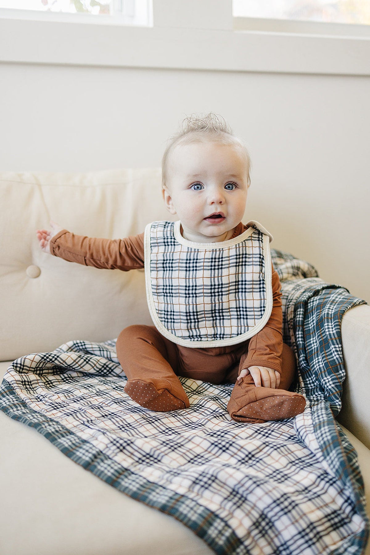 A baby wearing a vintage plaid muslin bib and brown outfit sits on a matching plaid blanket on a couch.