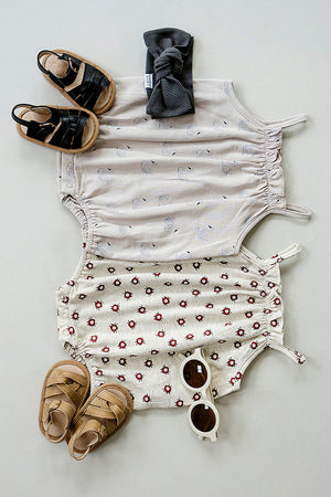 Two baby tank bubble rompers in cream with swan and floral patterns, paired with brown sandals and white sunglasses, arranged.