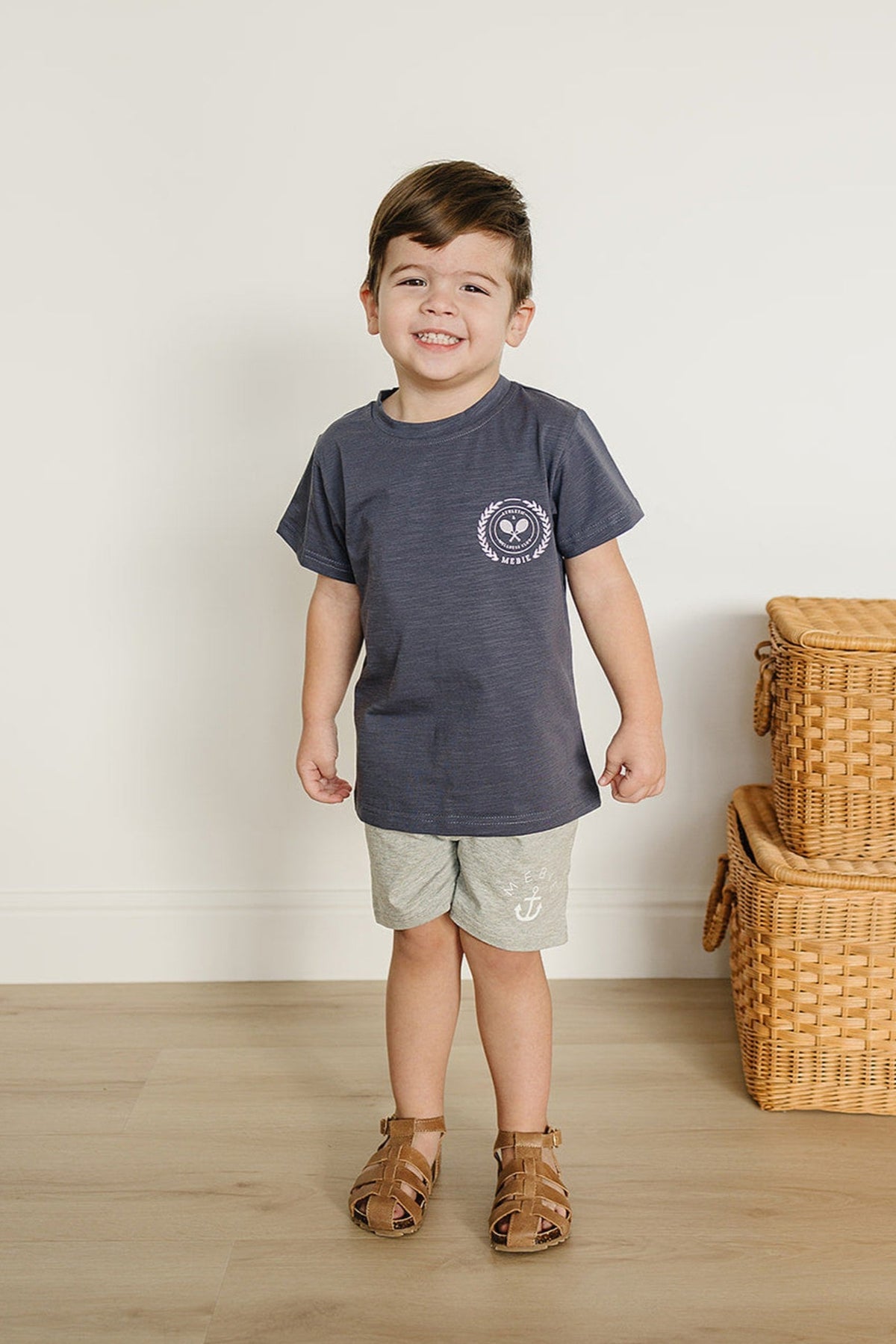 A child wearing heather grey shorts and a dark t-shirt stands smiling beside wicker baskets on a wooden floor.