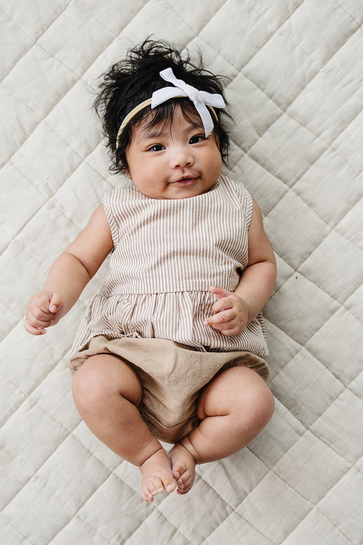A baby girl with dark hair wears a tan-striped peplum top and beige bloomers, lying on a quilted neutral blanket.