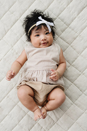 A baby girl with dark hair wears a tan-striped peplum top and beige bloomers, lying on a quilted neutral blanket.