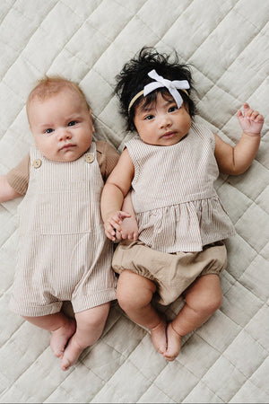 Two babies lie on a quilted beige surface, one wearing striped overalls and the other in a matching peplum top.