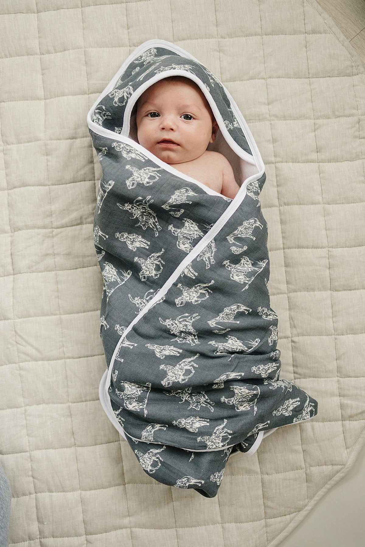 A baby is wrapped in a dark muslin hooded towel with a white rodeo pattern, lying on a quilted surface.