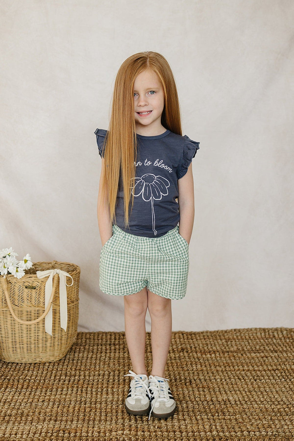 A child wearing green gingham shorts and a navy shirt with a floral print stands on a woven rug.