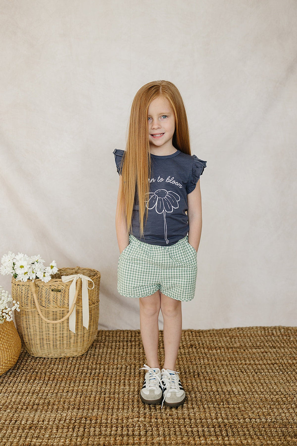 A young girl wearing a Born to Bloom navy ruffle t-shirt and green checkered shorts, standing on a woven rug.