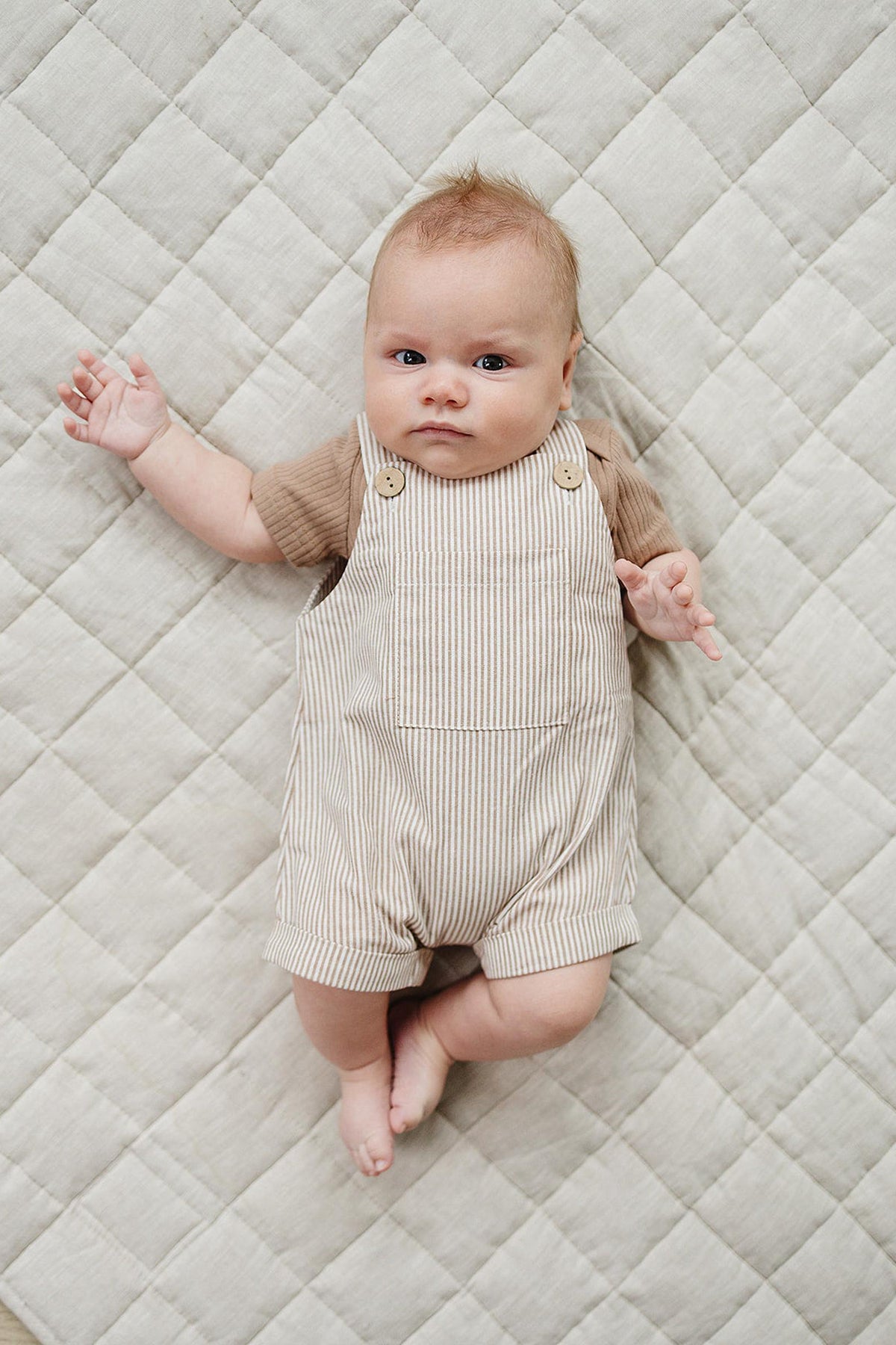 A baby lays on a quilted beige blanket, wearing a tan striped overall with a brown shirt underneath.