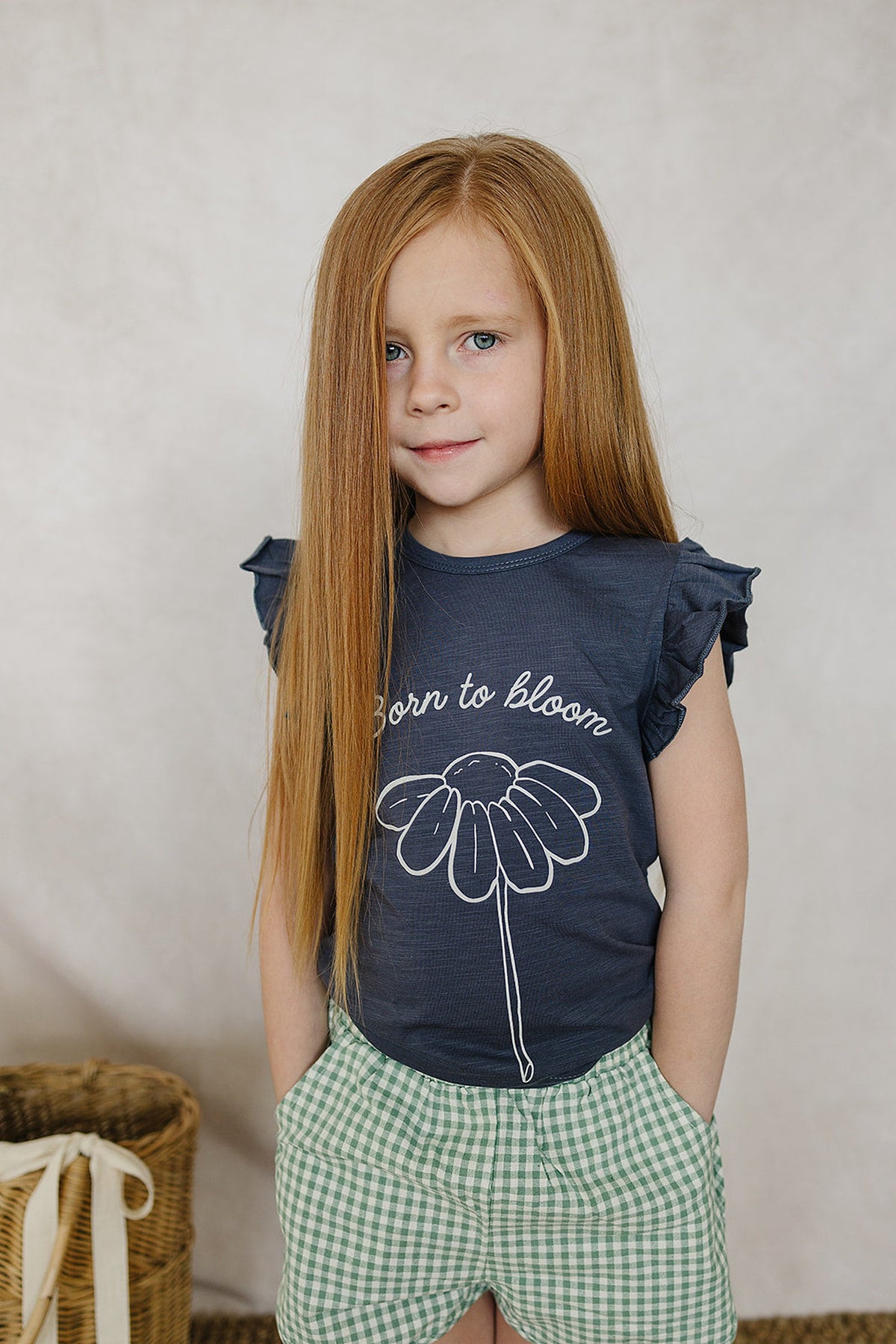 Young girl wearing a navy Born to Bloom ruffle t-shirt with a daisy graphic, paired with green checkered shorts.