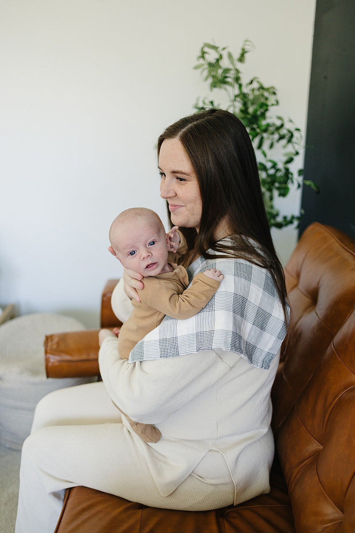 A woman holds a baby on her shoulder, using a plaid muslin burp cloth, seated on a brown sofa.