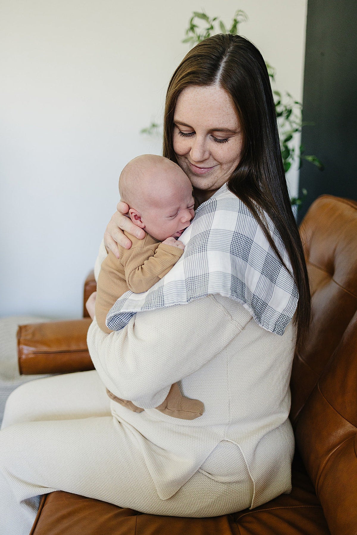 A woman holds a baby with a plaid muslin burp cloth on her shoulder, sitting on a brown couch.