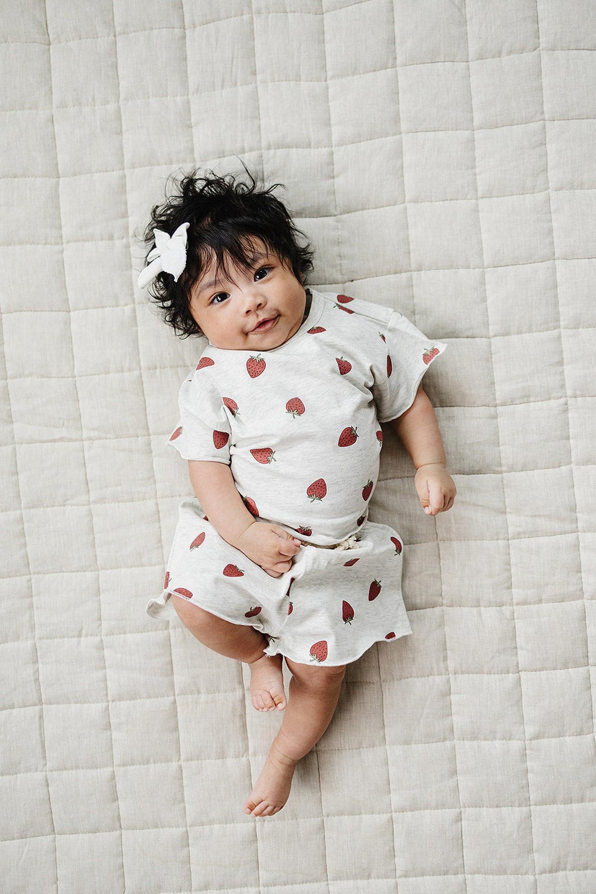A baby with curly hair wears a light grey cotton outfit featuring strawberry prints, lying on a textured beige blanket.
