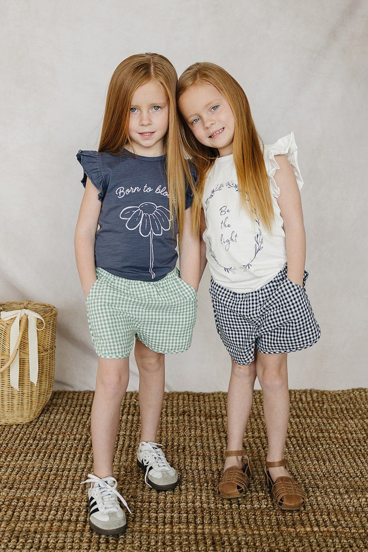 Two young girls wearing Born to bloom and Be the light ruffle sleeve shirts with gingham shorts, standing indoors.