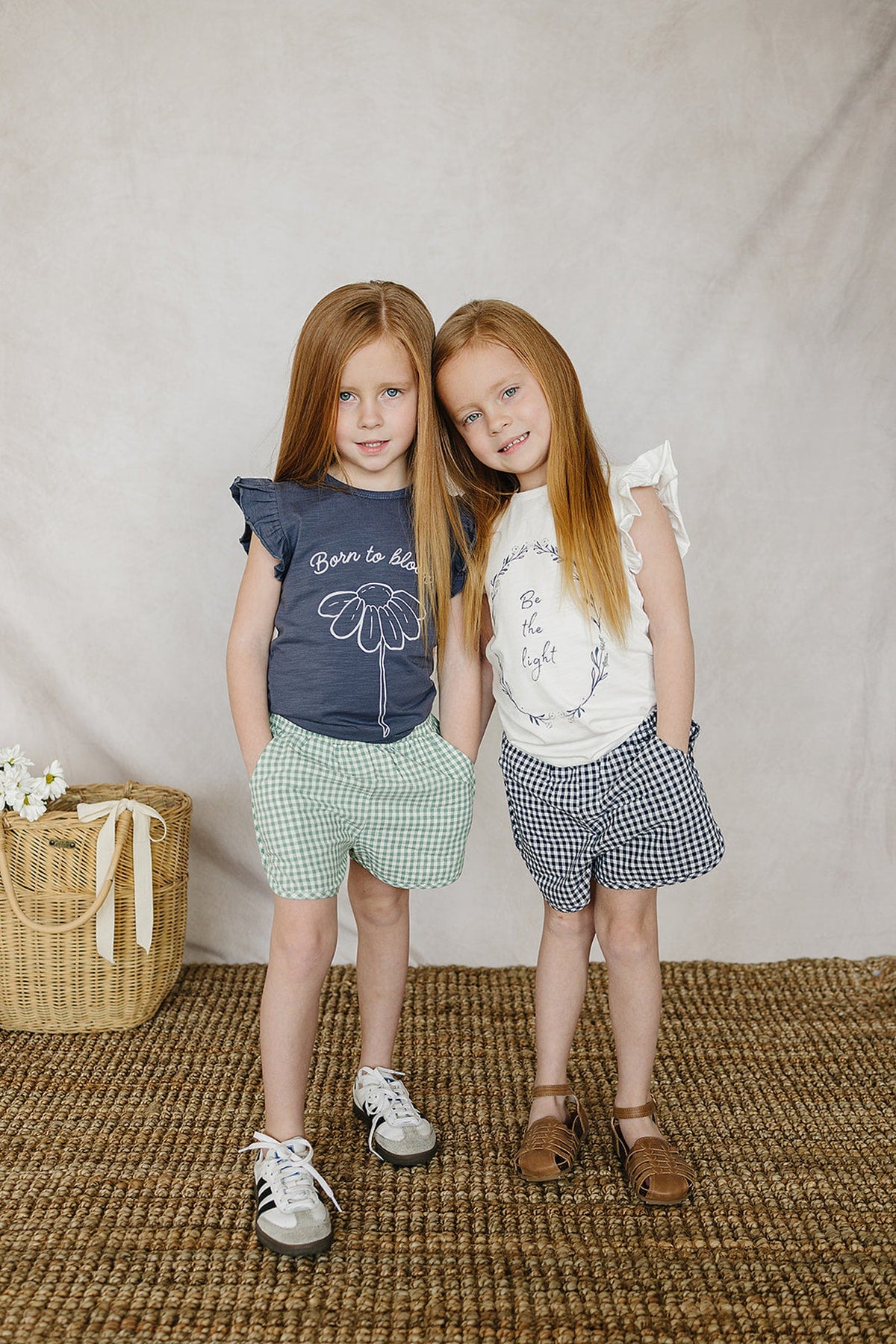 Two young girls in ruffle sleeve t-shirts and checkered shorts stand on a woven rug, smiling softly.