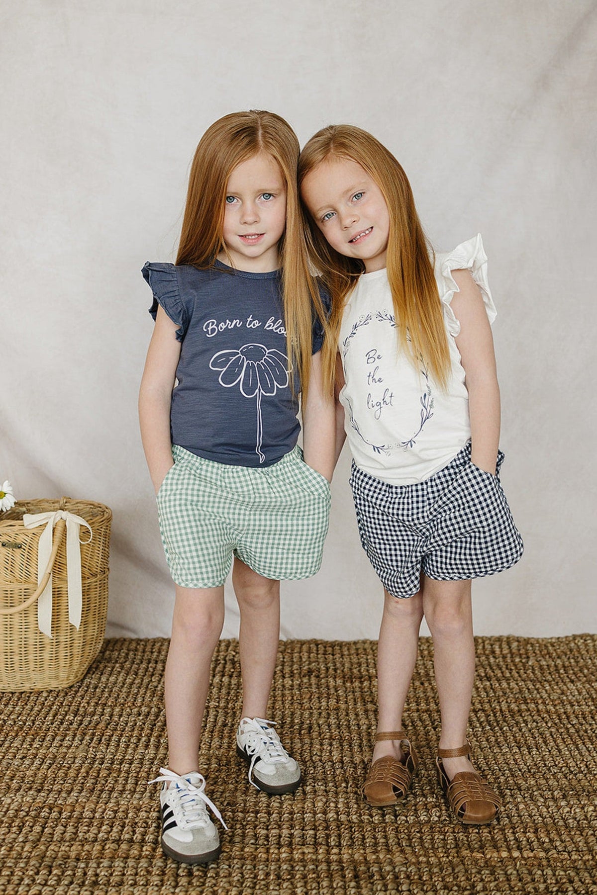 Two girls wearing gingham shorts, one in green and the other in blue, stand on a woven rug.