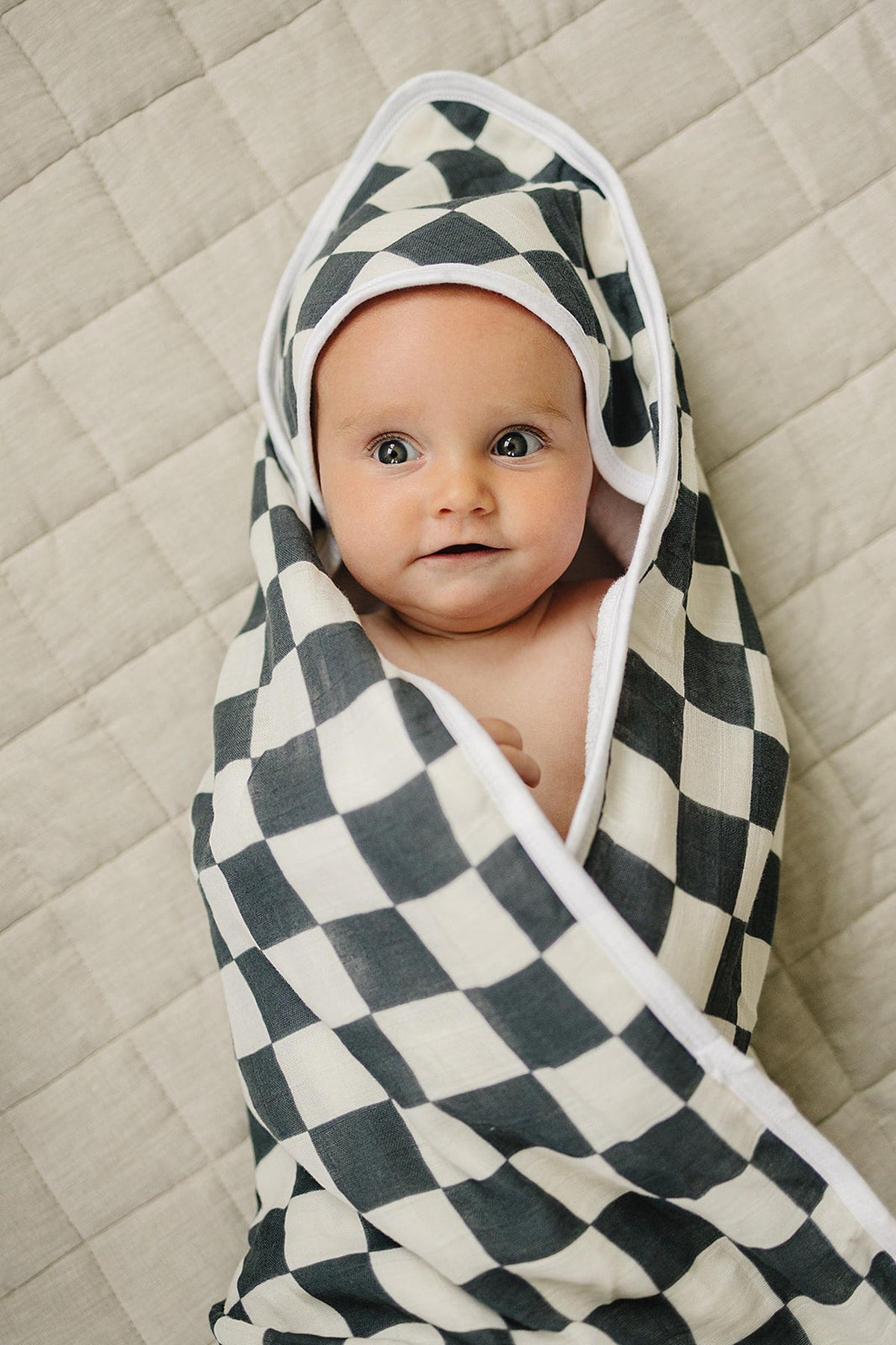 A baby wrapped in a charcoal checkered hooded towel, lying on a quilted beige surface, looking content.