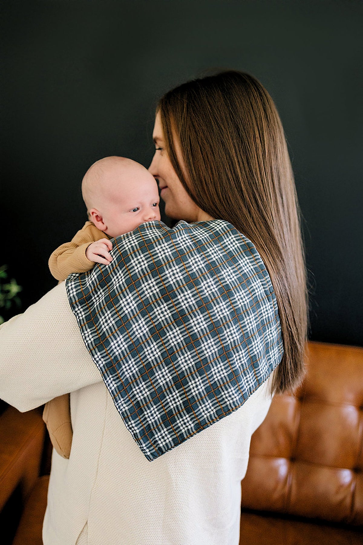 A woman with long hair holds a baby on her shoulder, showcasing a navy plaid muslin burp cloth made from 100% cotton.
