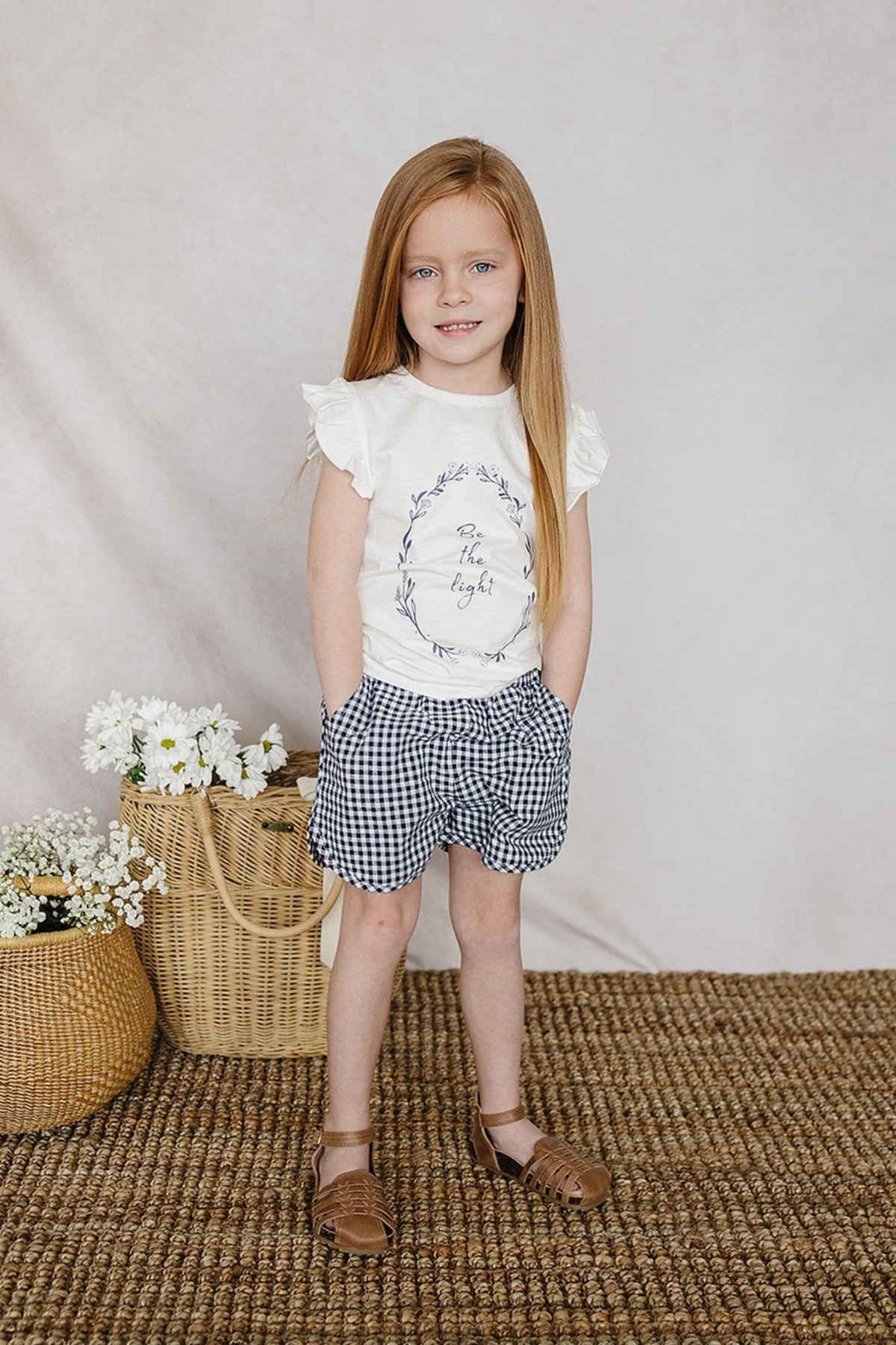 A child wearing a white Be the Light ruffle sleeve shirt and checkered shorts, standing on a woven rug.