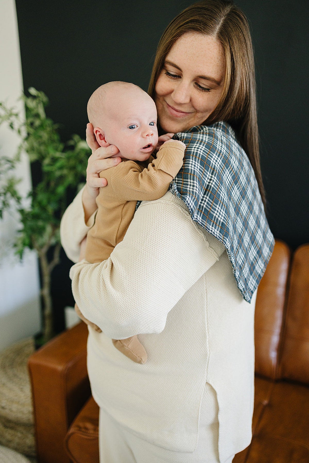 A mother holds a baby, showcasing a navy plaid muslin burp cloth draped on her shoulder. The cozy setting features a brown leather couch and green plants in the background.