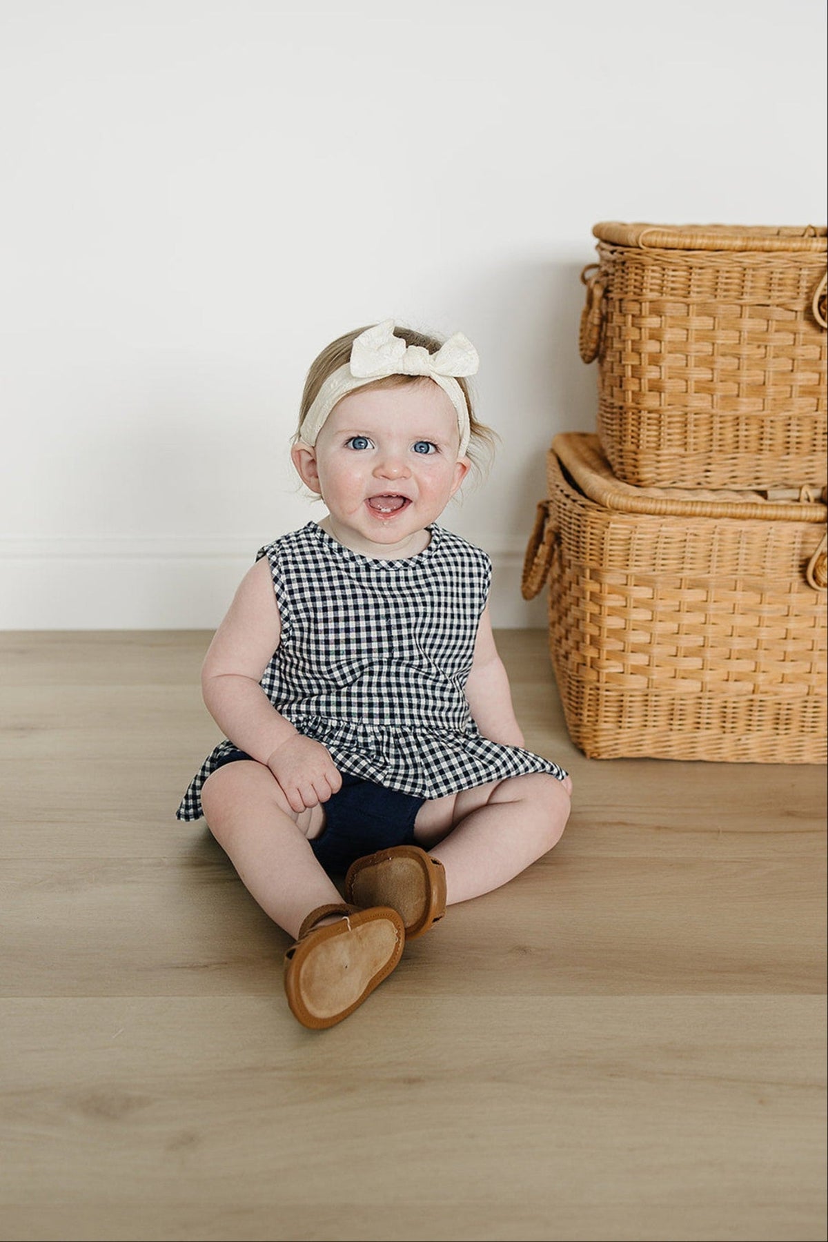 A baby in a navy gingham peplum set sits smiling on a wooden floor near wicker baskets.