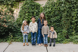 Five children stand together outdoors in front of a lush green wall, each wearing cozy sweaters and casual jeans, showcasing a playful and stylish atmosphere perfect for family gatherings.