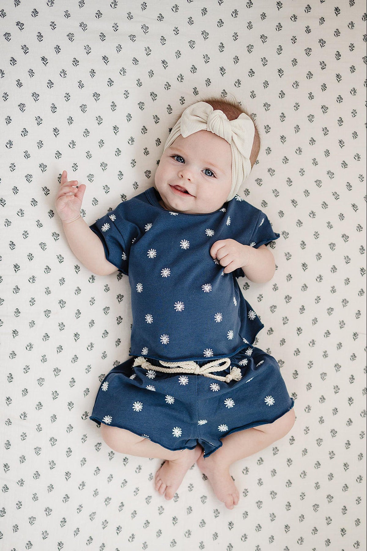 Baby wearing a navy daisy-print cotton set with raw hem, lying on a floral-patterned sheet, smiling.