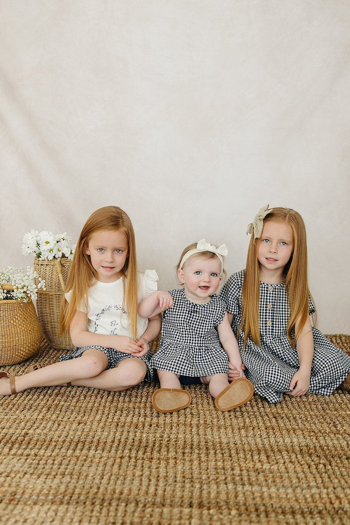 Three young girls sit on a textured rug, wearing coordinated gingham dresses. A woven basket with flowers is nearby.