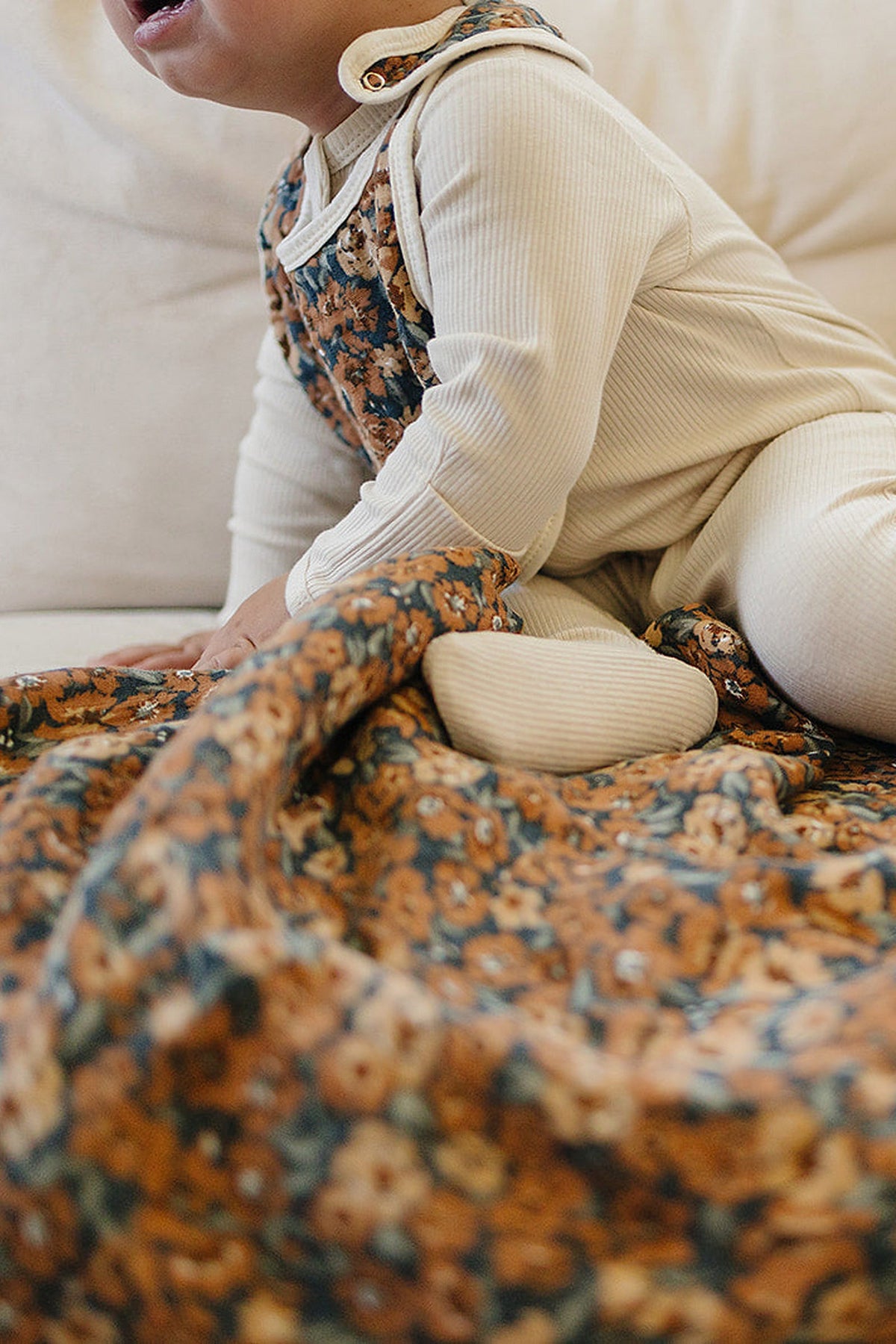 A baby wearing a cream outfit sits on a floral-patterned quilt with blue and orange flowers.