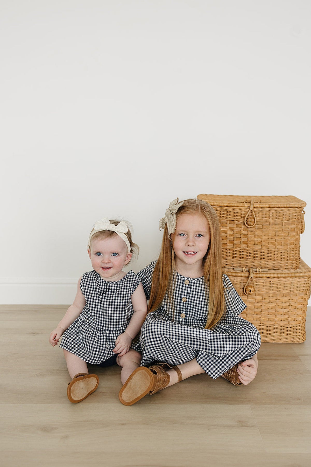 Two young girls in navy gingham outfits sit on a wooden floor, with wicker baskets in the background.