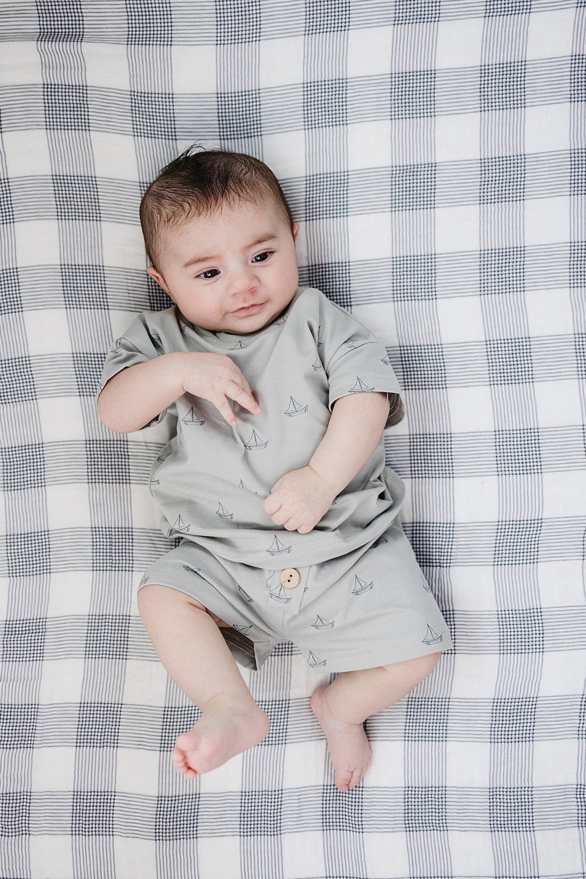A baby lies on a checkered blanket, wearing a light grey short-sleeve top and matching shorts adorned with a subtle sailboat pattern. The outfit is made from soft, breathable cotton.