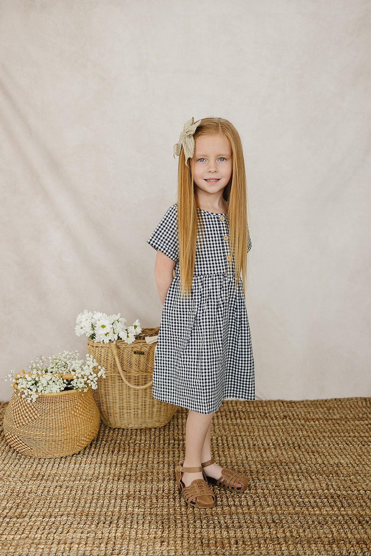A young girl smiles while wearing a navy gingham cotton dress, paired with brown sandals, in a softly lit setting.