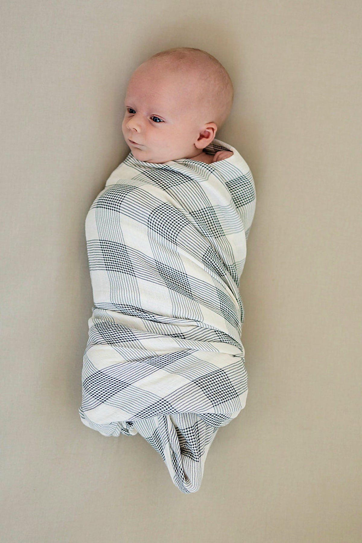 A baby is swaddled in a black and white plaid muslin blanket on a neutral background.
