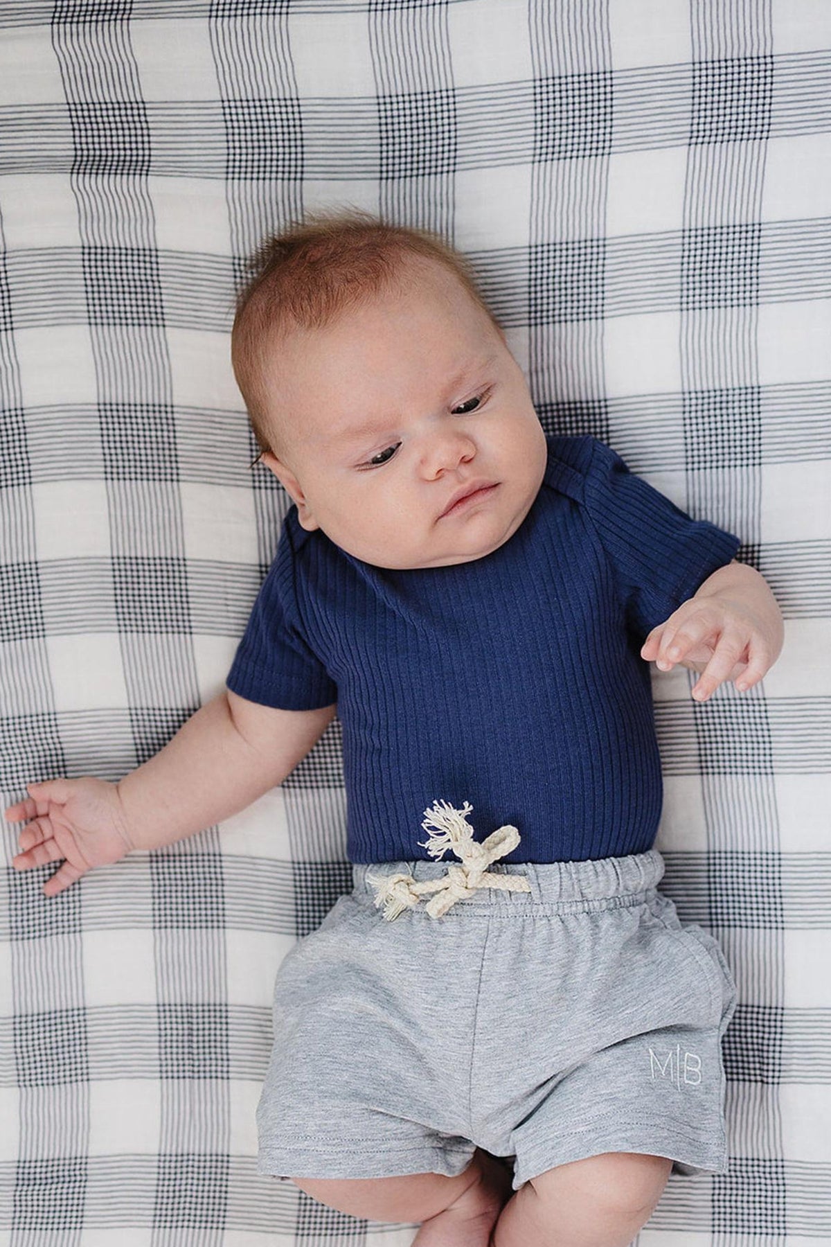A baby lies on a checkered blanket, wearing a navy ribbed bodysuit and gray shorts with a decorative tie.