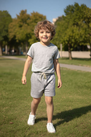 A young boy smiles while walking in a park, wearing grey cotton pocket shorts and a matching grey t-shirt. The outfit is comfortable and perfect for outdoor play.