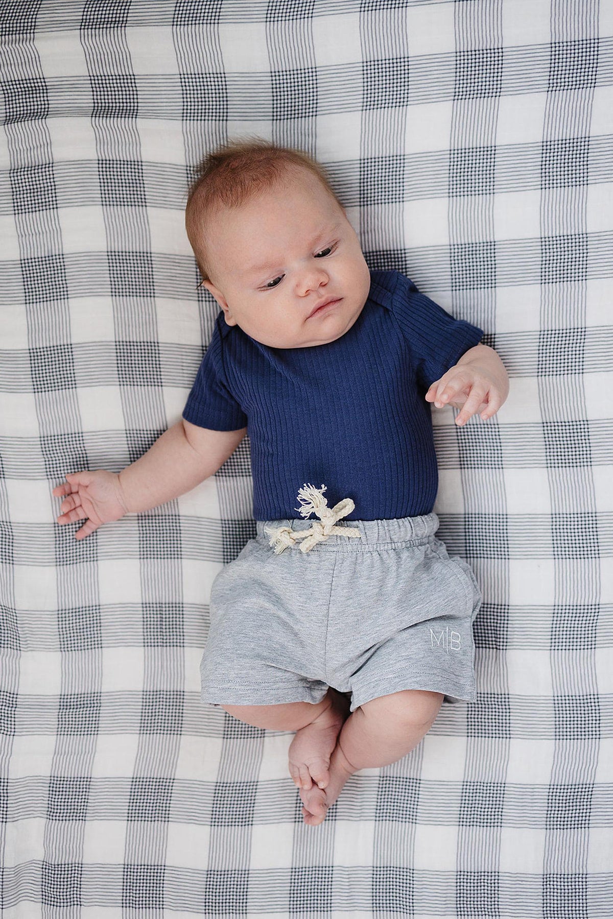 A baby lies on a checkered blanket, wearing a navy ribbed bodysuit and soft grey cotton shorts with a decorative drawstring.