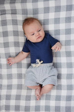 A baby lies on a checkered blanket, wearing a navy ribbed bodysuit and soft grey cotton shorts with a decorative drawstring.