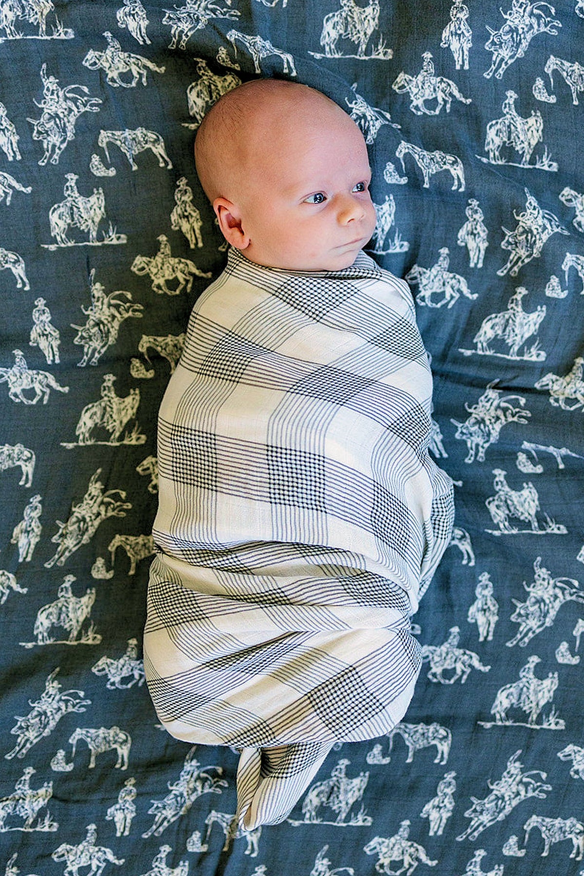 A baby is wrapped in a black and white plaid swaddle blanket, lying on a cowboy-patterned fabric.