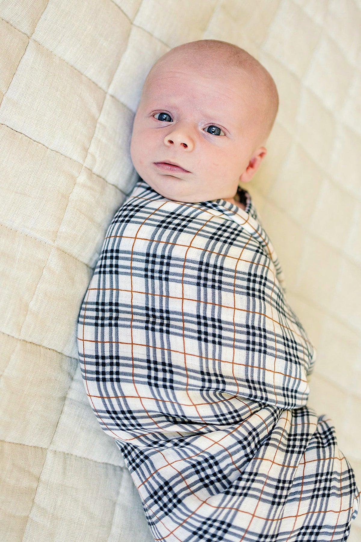 A baby wrapped in a vintage plaid muslin swaddle blanket rests on a quilted cream surface.