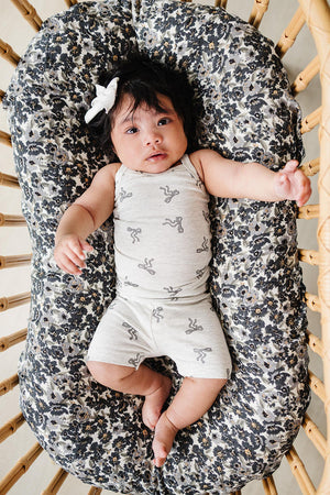 Baby in light gray bow-patterned tank top and shorts lying on a floral cushion in a wooden crib.