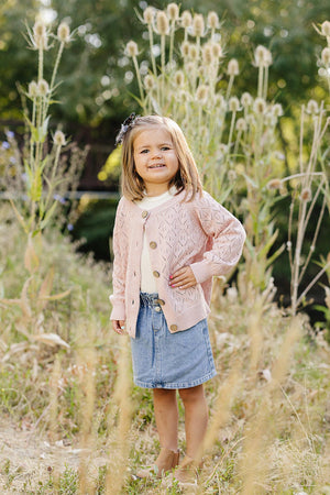 A young girl stands in a field wearing a pink button-down cardigan and denim skirt, smiling warmly.